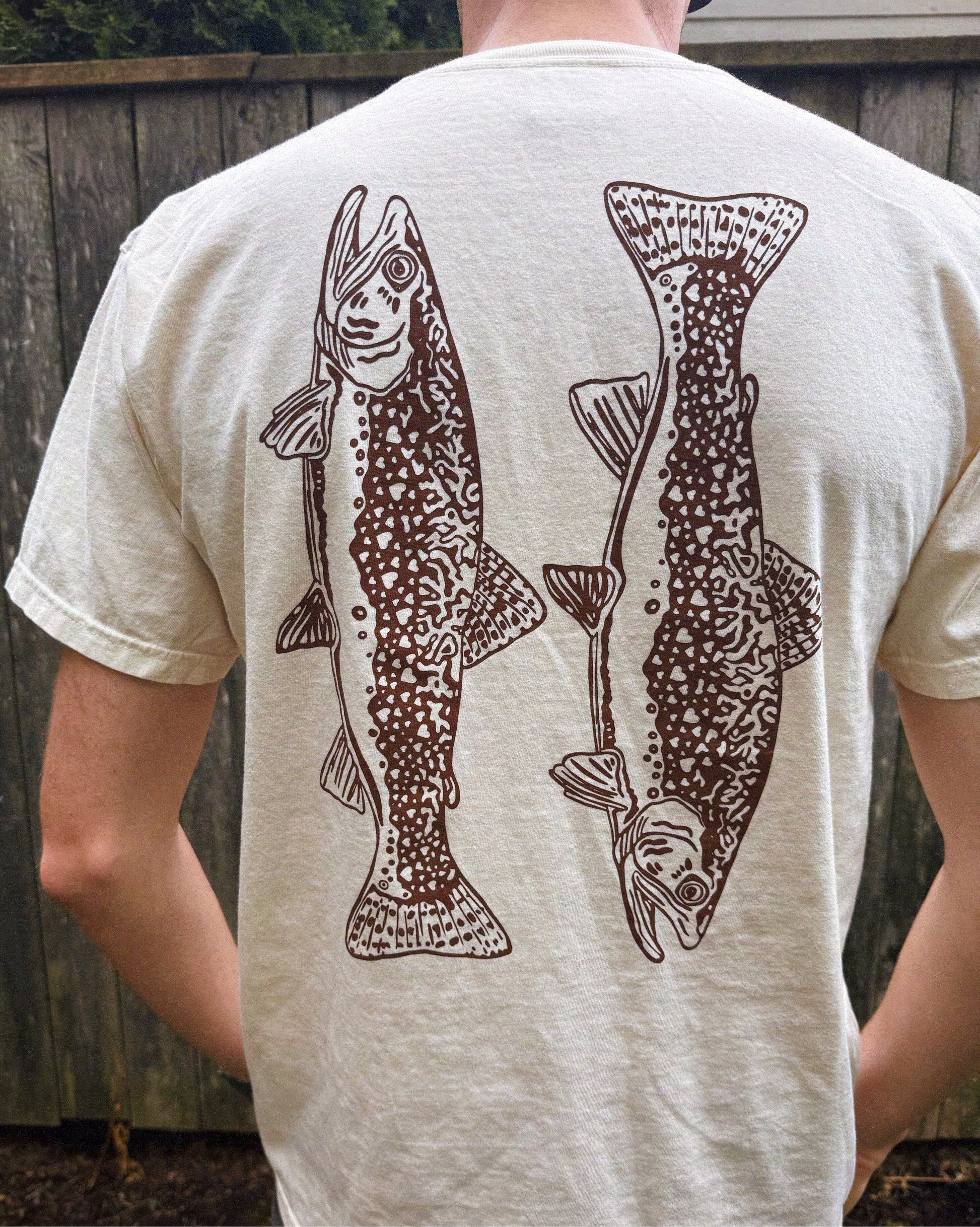 Person wearing a beige t-shirt with brown fish illustrations on the back.