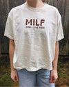 Person wearing a white t-shirt with 'MILF (Man I Love Fish)' text in front of a wooden fence.