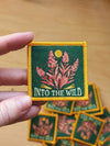 Embroidered patch with 'Into the Wild' text and floral design held by a hand on a wooden surface.